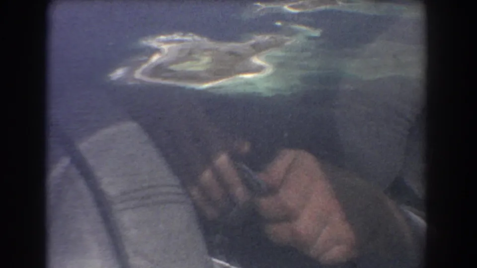 1967 travel footage frame showing a small island ringed by pale water, filmed past a passenger hand from an airplane window.
