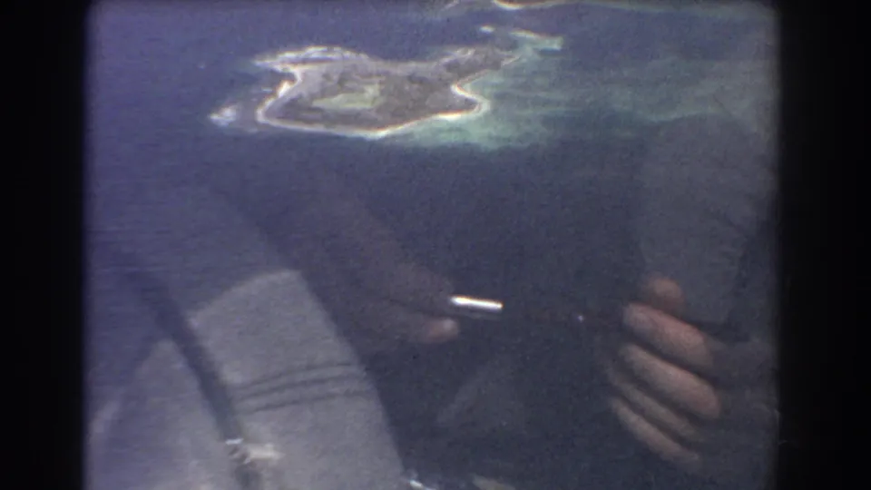 A small island and lagoon remain centered outside the airplane window, while passenger hands and cabin shadows appear below.