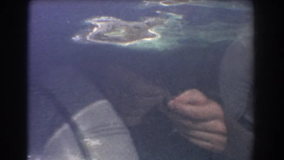 1967 8mm travel frame from Perth, Australia metadata showing a small island below and a woman's reflected hands in the airplane window.