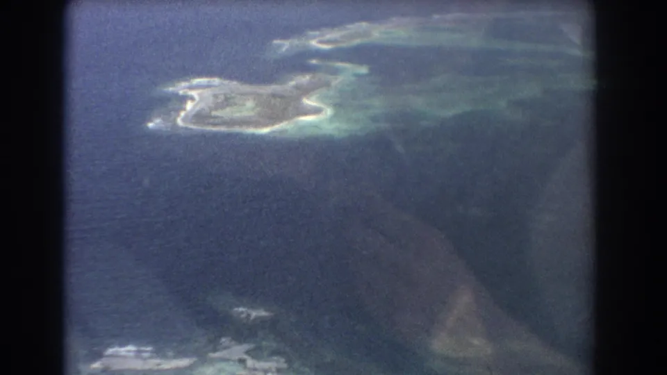 Soft, grainy 8mm aerial view of a small coastal island and turquoise shallows, with faint airplane-window reflection across the frame.