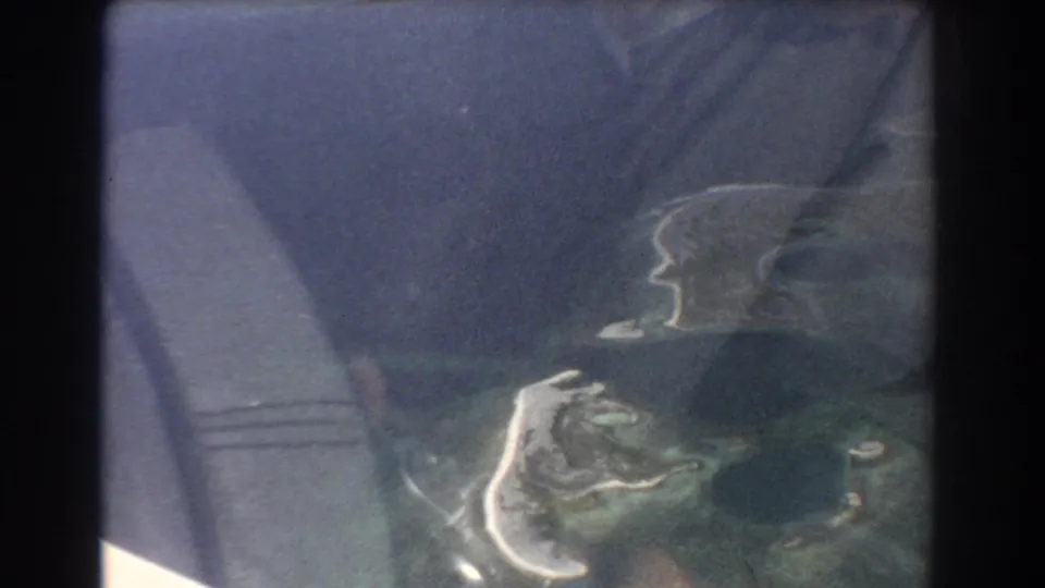 Window-reflected airplane interior and hands overlay a partial island view in this 1967 Perth, Australia travel frame.