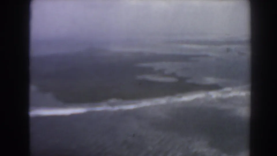 1967 8mm aerial view near Perth, with a dark coastal shape, white breakers, and open sea under haze.