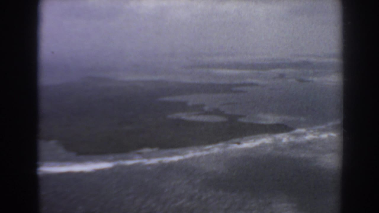 Aerial view of surf breaking along a dark coastline through aircraft-window framing.