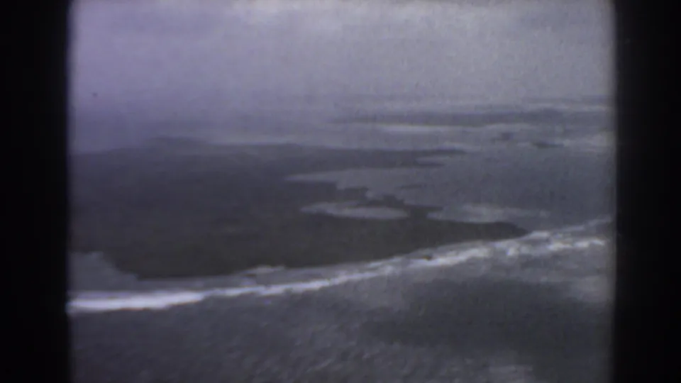1967 8mm aerial view near Perth, Australia, showing ocean surf, low shoreline, and dark aircraft-window edges.
