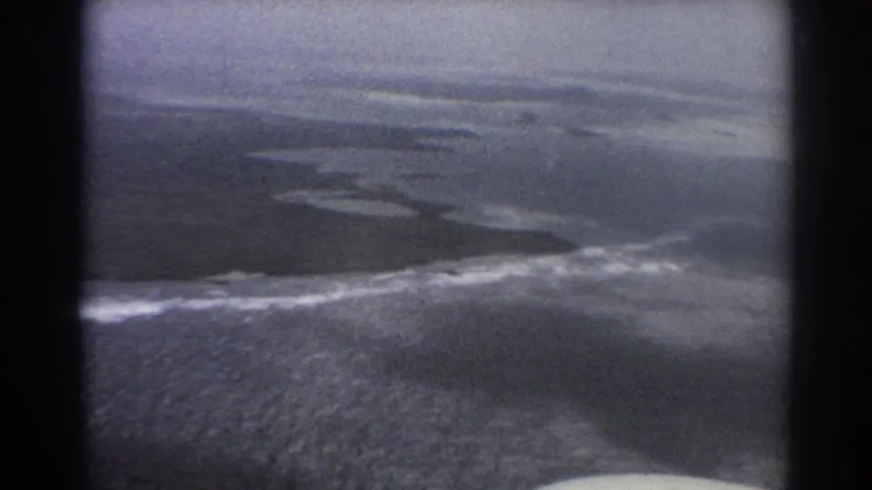 Aerial 8mm frame near Perth in 1967 with ocean, white surf, low coastal land, and heavy dark framing from the aircraft window.