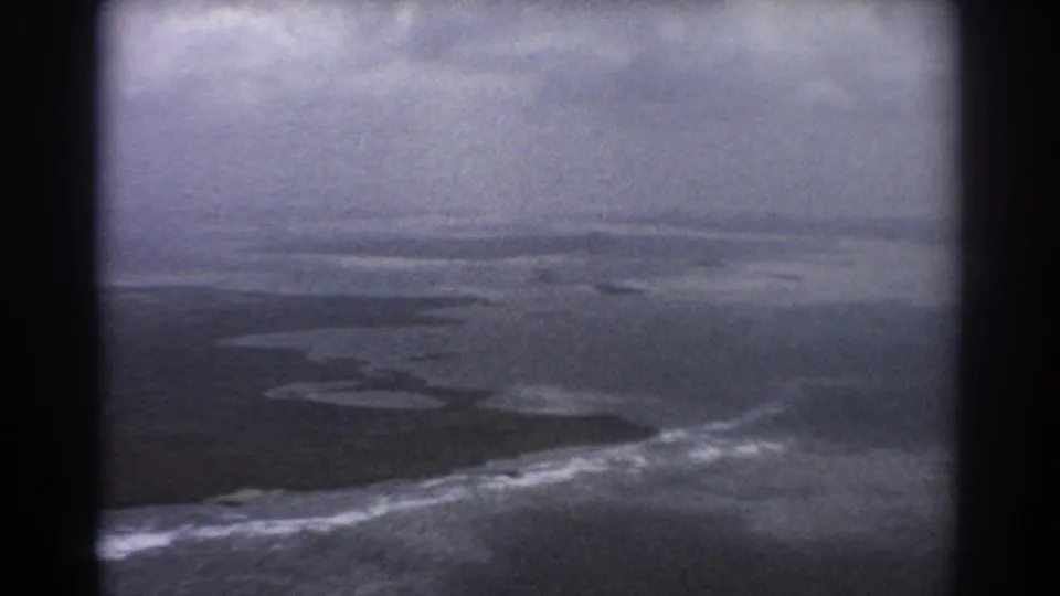 1967 home-movie aerial frame near Perth showing broad sea, surf bands, dark coastal land, and hazy sky in grainy 8mm texture.