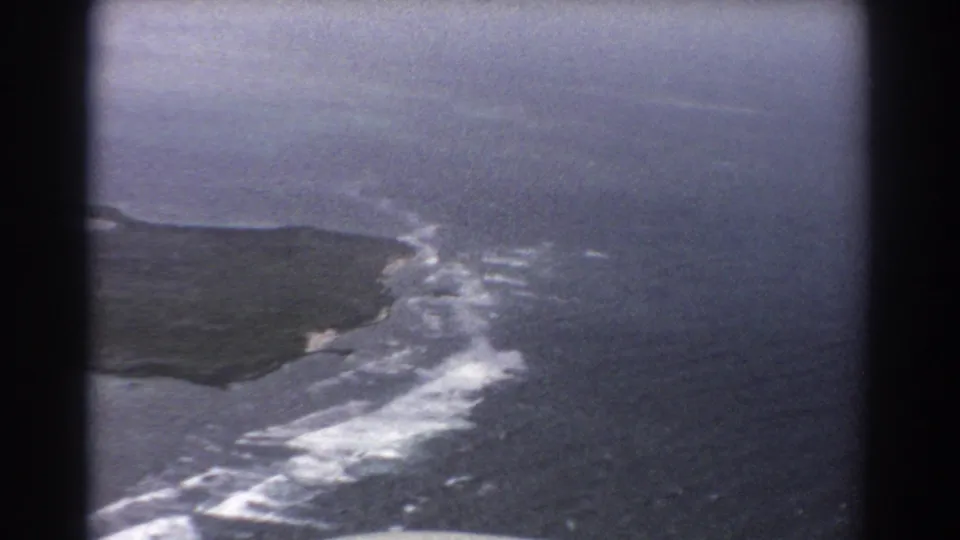 Near Perth in 1967, the 8mm frame shows a coastal headland, white breaking waves, open water, and soft aircraft-window framing.