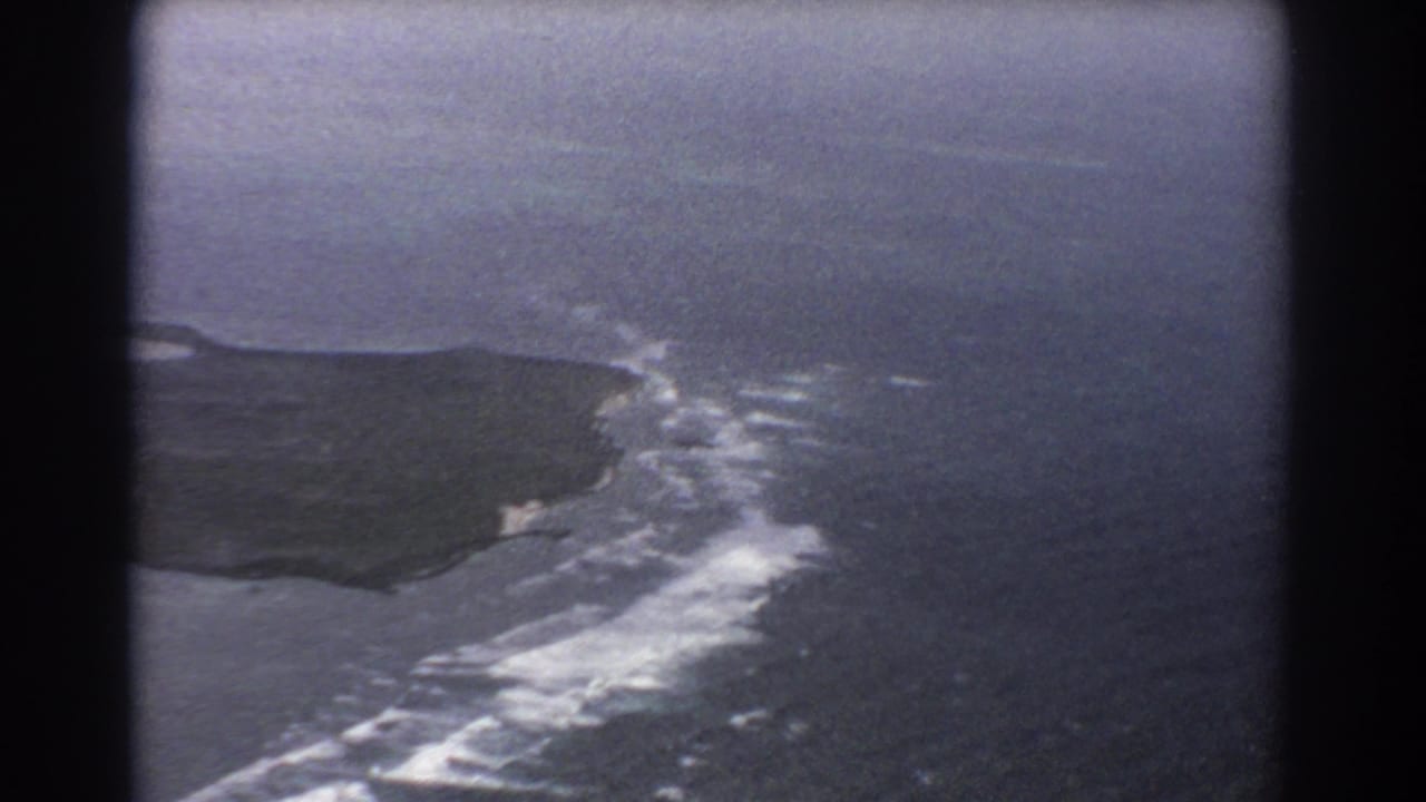 Aerial view of a dark island coastline with white surf and blue-gray ocean.