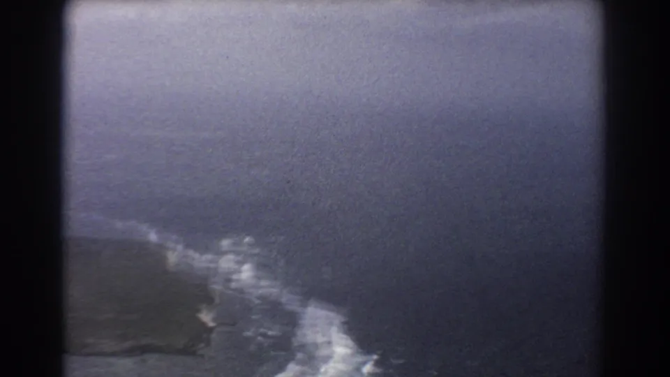 Muted 1967 8mm view near Perth showing white waves along a coast under haze, with the aircraft perspective softened by film grain.