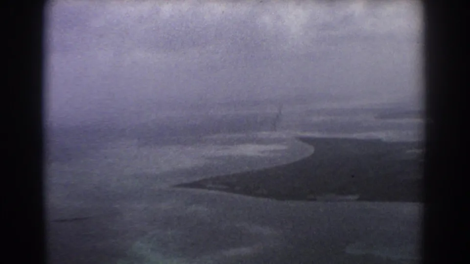 1967 8mm airplane-window view near Perth showing water, distant coastline, clouded sky, and a dark aircraft wing or window edge.