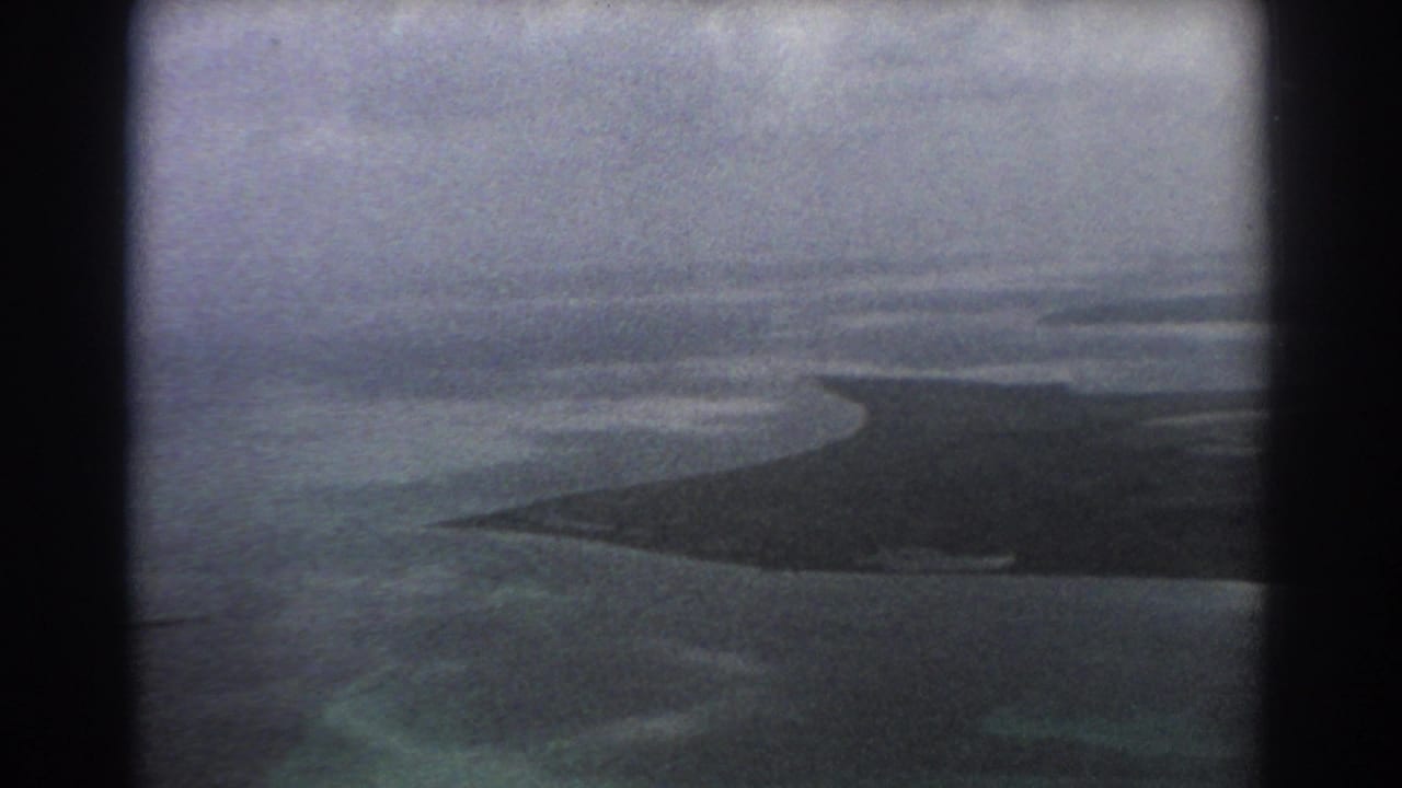 1967 Perth, Australia 8mm aerial frame showing a dark low island or sandbar in blue-gray coastal water, seen through aircraft-window edges.