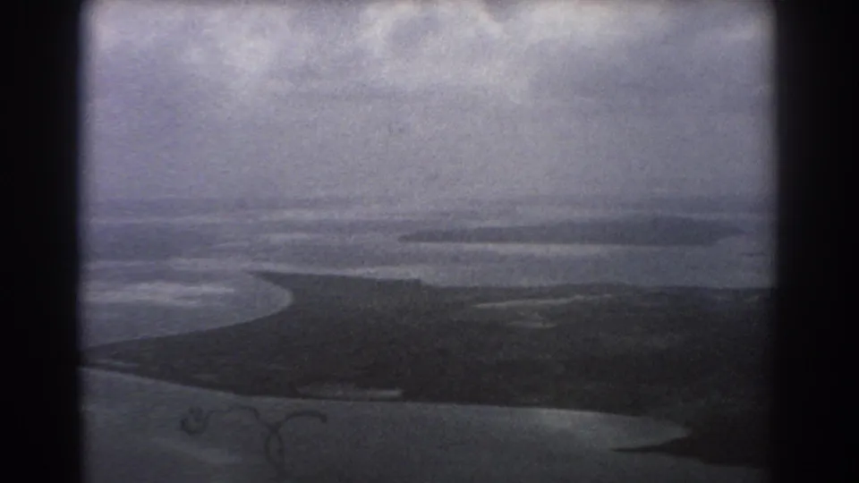 1967 Perth, Australia aerial home-movie frame with muted water, a low island or sandbar, cloudy sky, and dark film gate or window edges.
