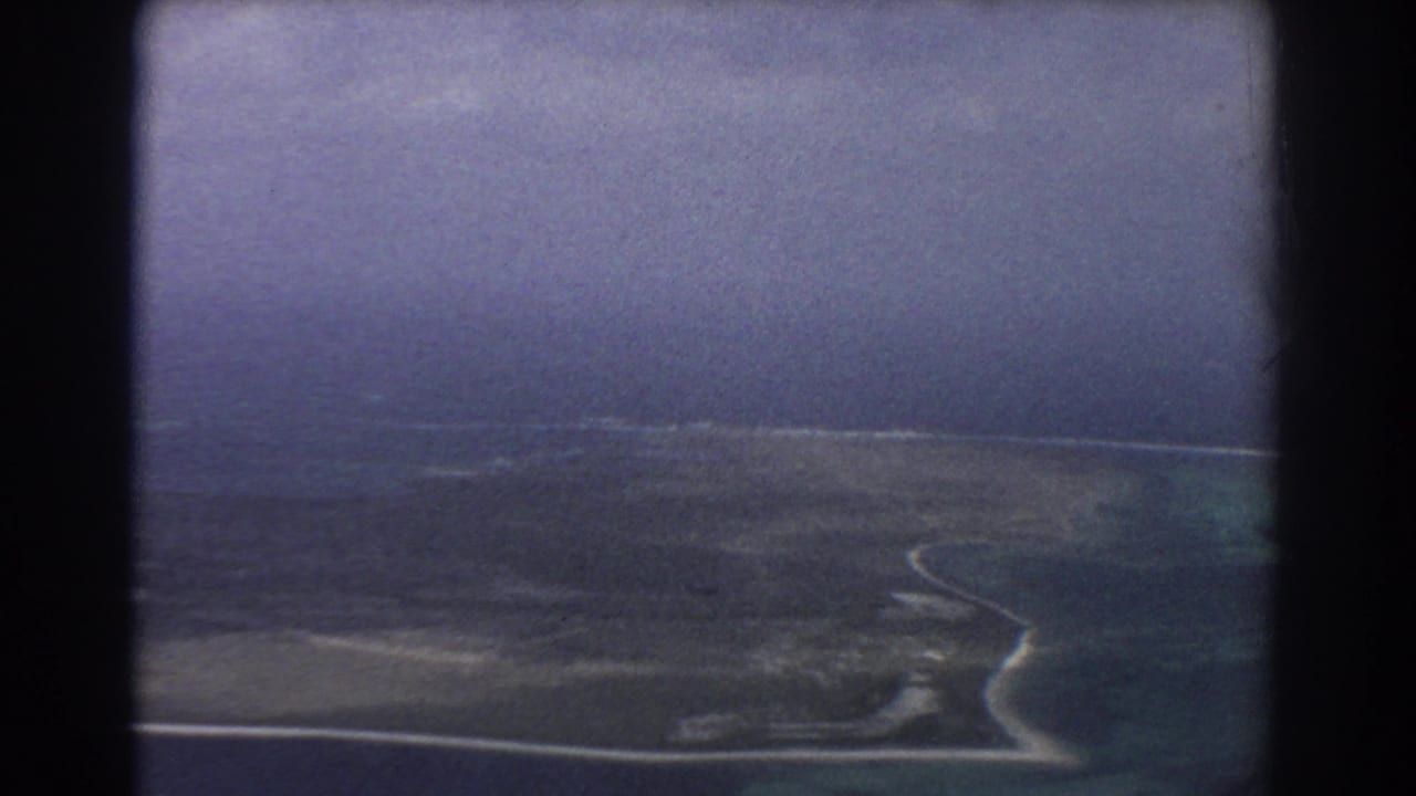 Aerial view of dark coastal land, pale shoreline, and cloudy sky through a film frame.