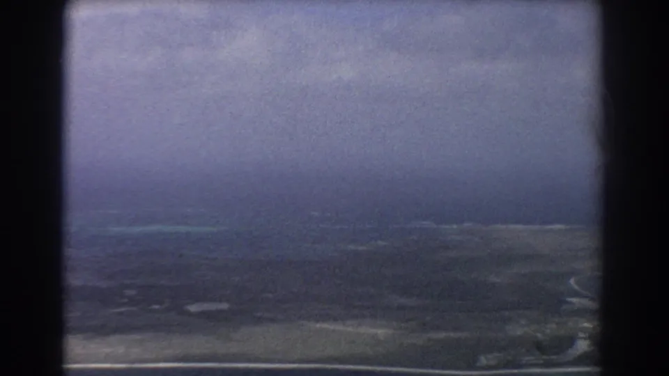 Aerial 8mm frame of coastal Western Australia, with low land, pale beach line, shallow turquoise water, and overcast sky.