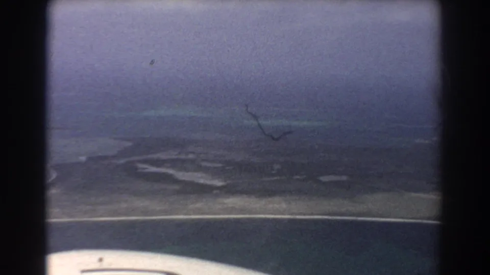 1967 Perth-area aerial frame with turquoise shallows, pale coast, dark terrain, cloudy sky, and a partial aircraft wing.