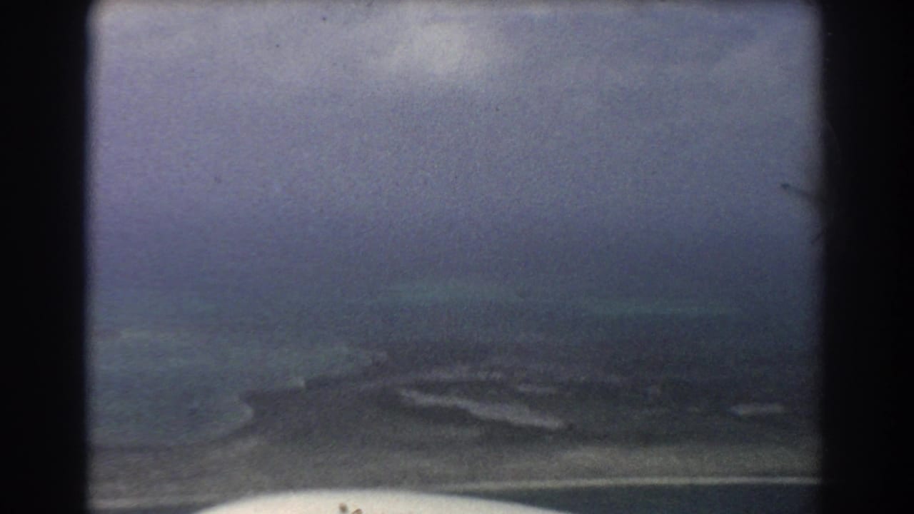 Aerial view through a dark aircraft window frame over blue-green water and a low coastal landform.