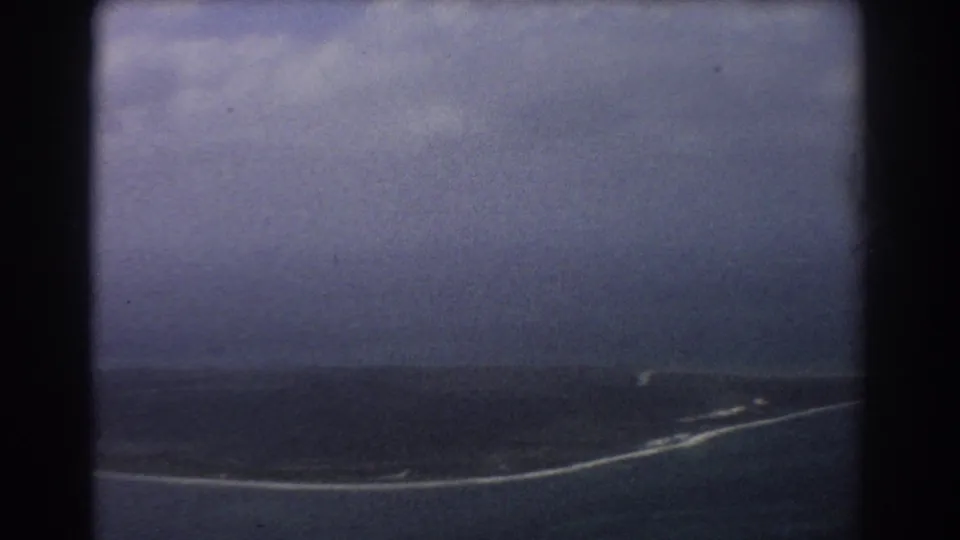 1967 home-movie aerial near Perth, Australia, with a narrow island-like strip, surf line, and overcast sky seen from a plane.