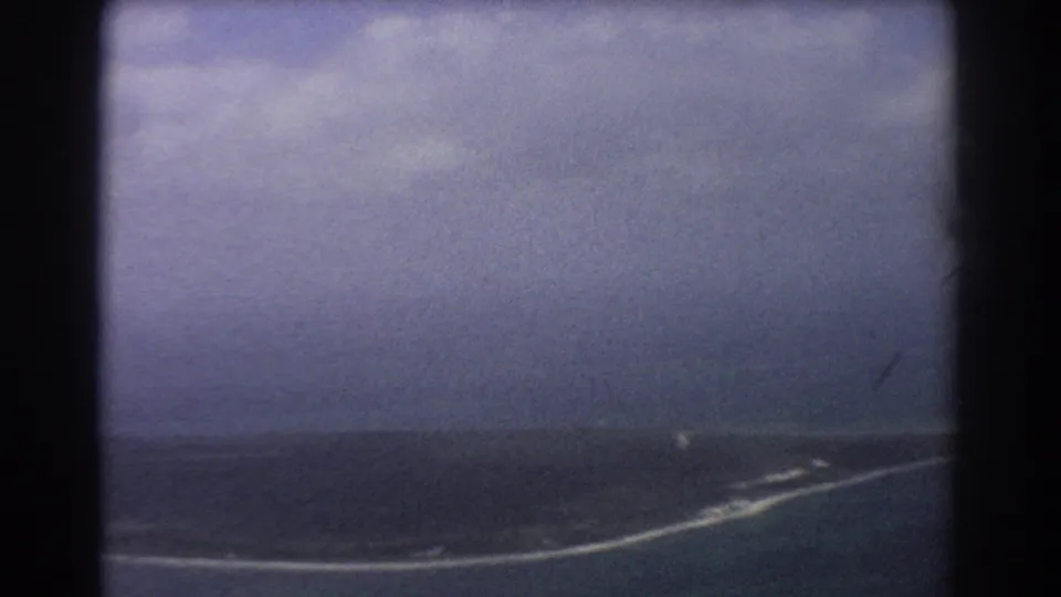 1967 8mm airplane-window view near Perth, Australia, showing a low coast or island edge with pale surf and subdued ocean color.