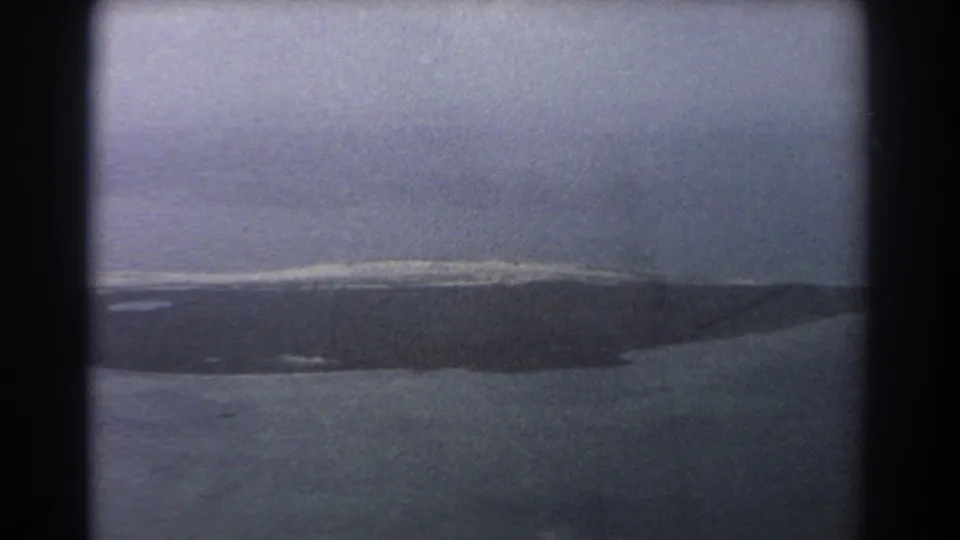 1967 8mm aerial frame near Perth with an elongated island shape, soft surf edges, calm sea, and muted blue cast.