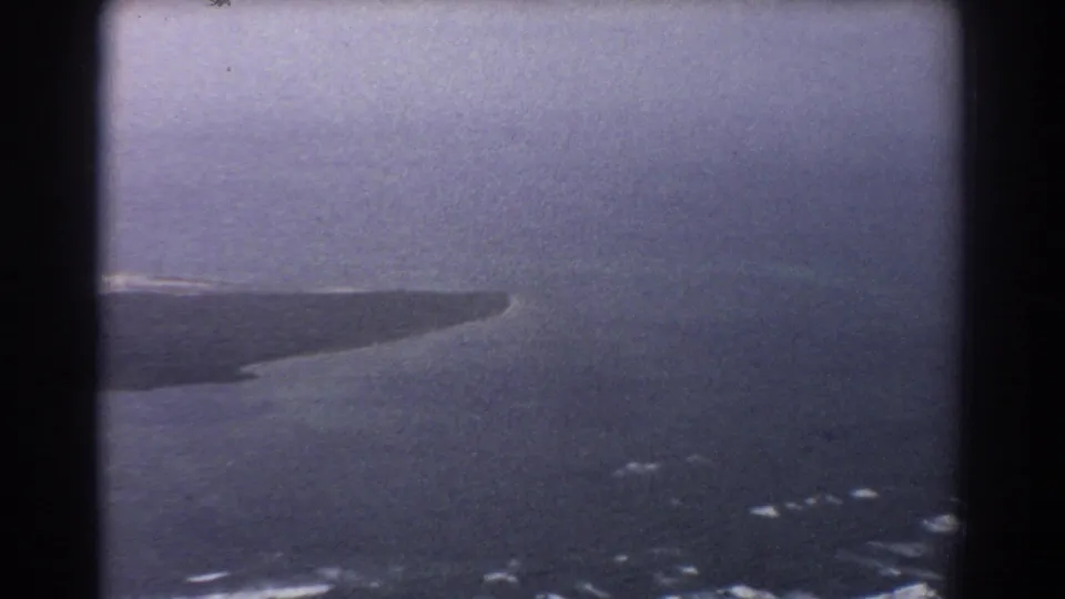 1967 8mm aerial frame near Perth with a distant island edge, broad ocean surface, scattered surf, and visible film grain.