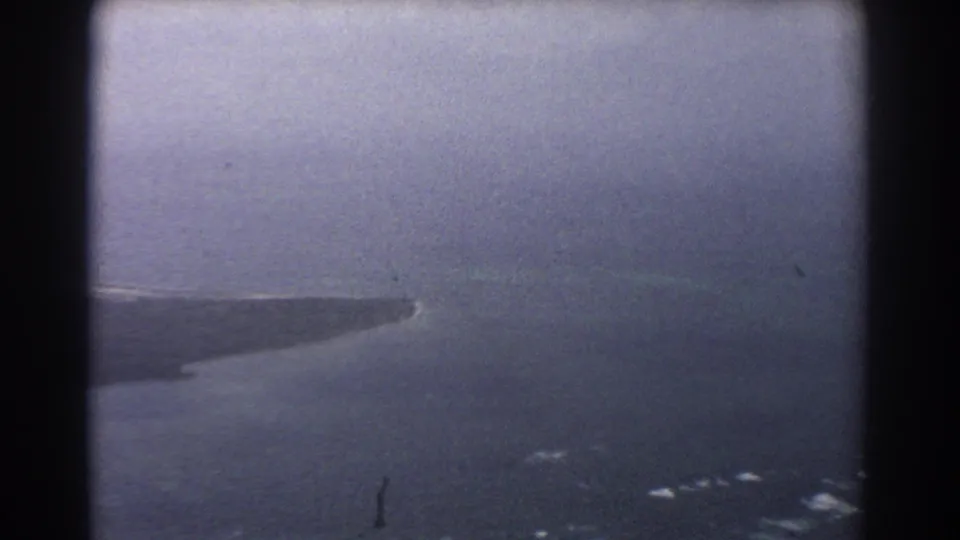 1967 Perth-area 8mm aerial view of a low barren coastal landform surrounded by dark water and haze.