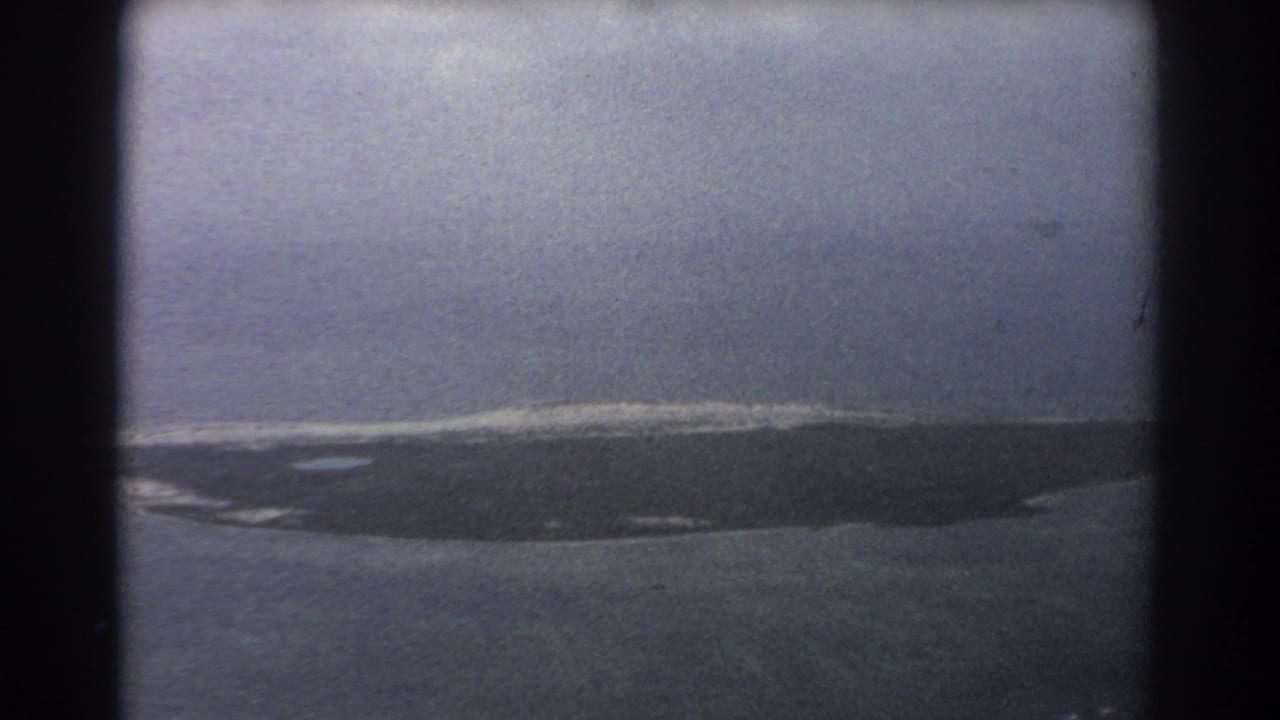 Aerial view of a low island and surrounding ocean through an airplane window.