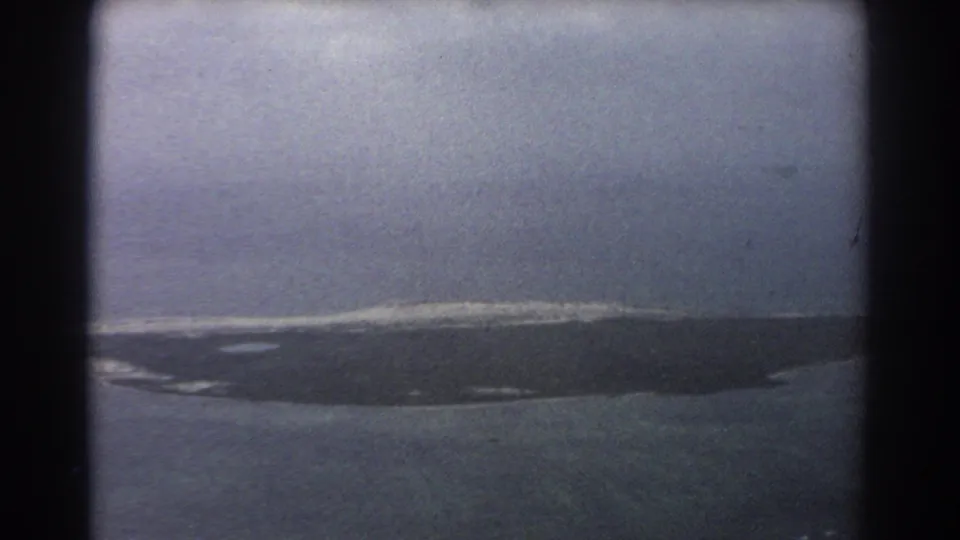 1967 8mm-style aerial view near the Perth, Australia metadata context, showing a dark island edge, pale reef line, and ocean through an aircraft window.