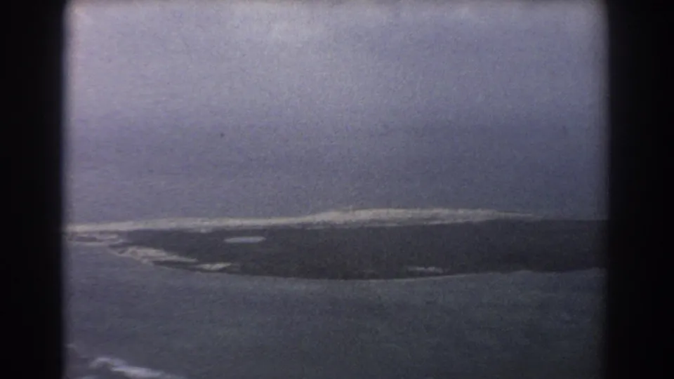 Aerial home-movie frame showing island shoreline, surf or reef edge, dark water, and heavy aircraft-window borders in the 1967 travel footage.