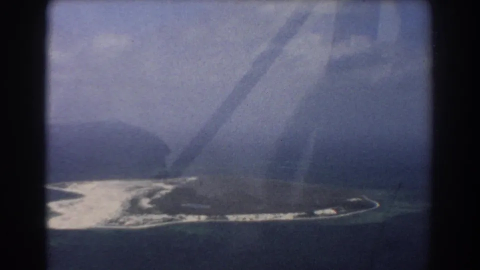 Aerial view of a pale sandbar, curved island edge, and surrounding water, partially obscured by airplane-window reflections in 1967 8mm footage.