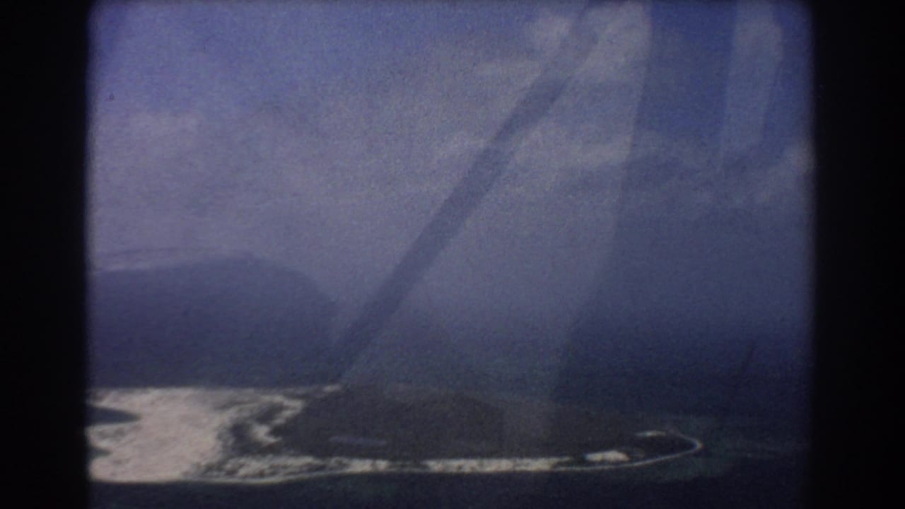 Aerial view of a small island and pale surf line seen through a dark airplane window frame.
