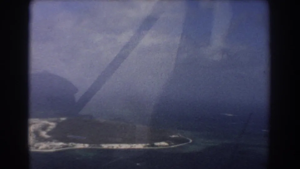 1967 8mm home-movie frame of an island coastline from above, with surf, blue-green water, haze, and dark aircraft-window structure.