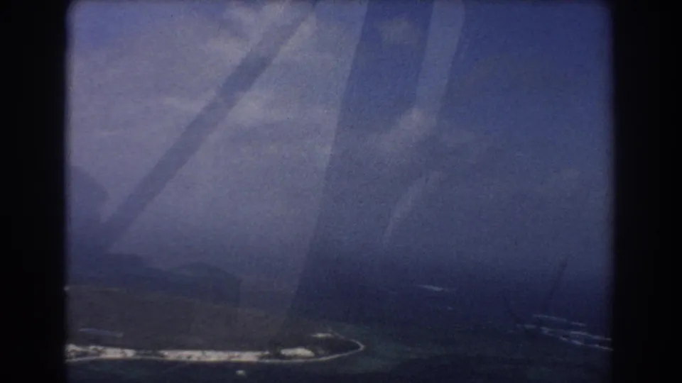 1967 8mm aerial footage frame with a hazy island edge, surrounding water, film grain, and prominent aircraft-window shadows.