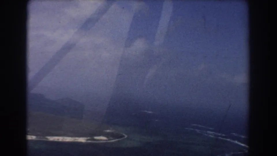 1967 8mm aerial view near Perth metadata context, with island shoreline, turquoise water, soft haze, and dark window-frame shadows.