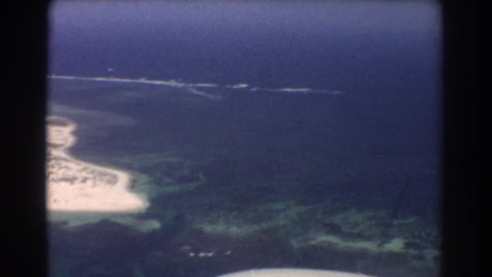 Aerial 8mm view over Perth, Australia coastline with pale beach, reef-colored shallows, and dark aircraft window edges.
