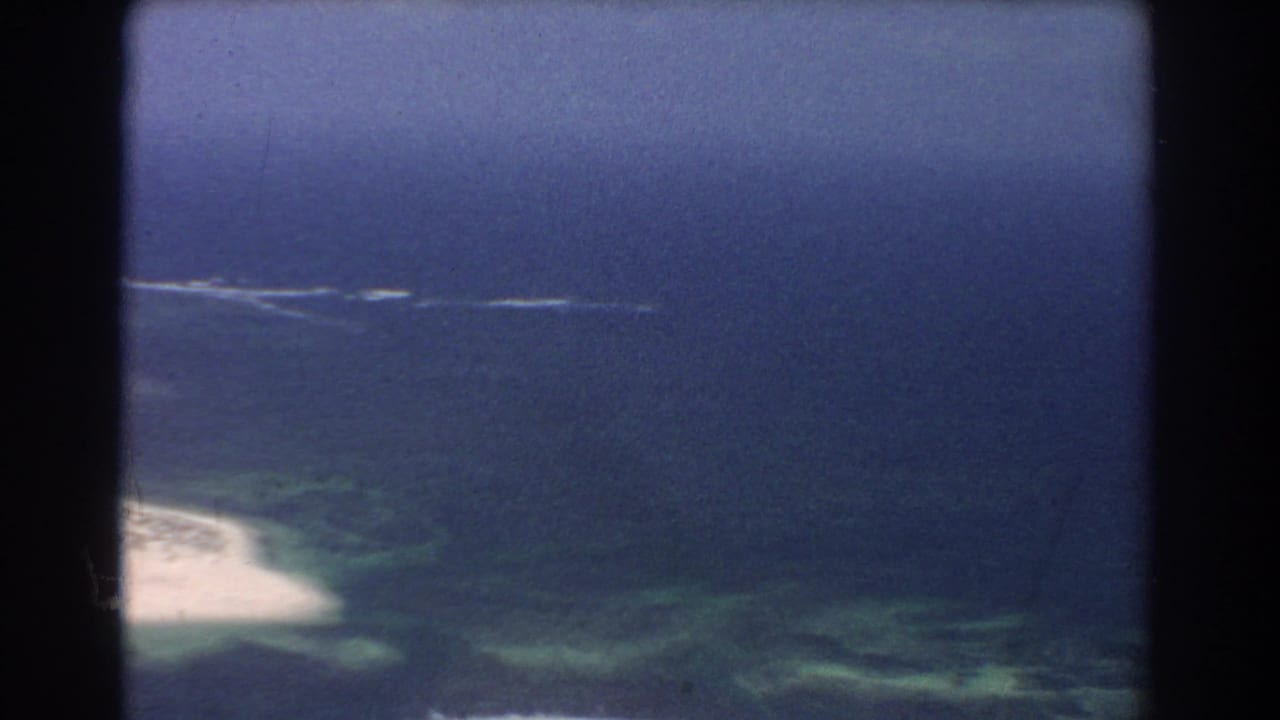 Aerial view of blue ocean, pale sand, and green shallows through dark 8mm frame borders.