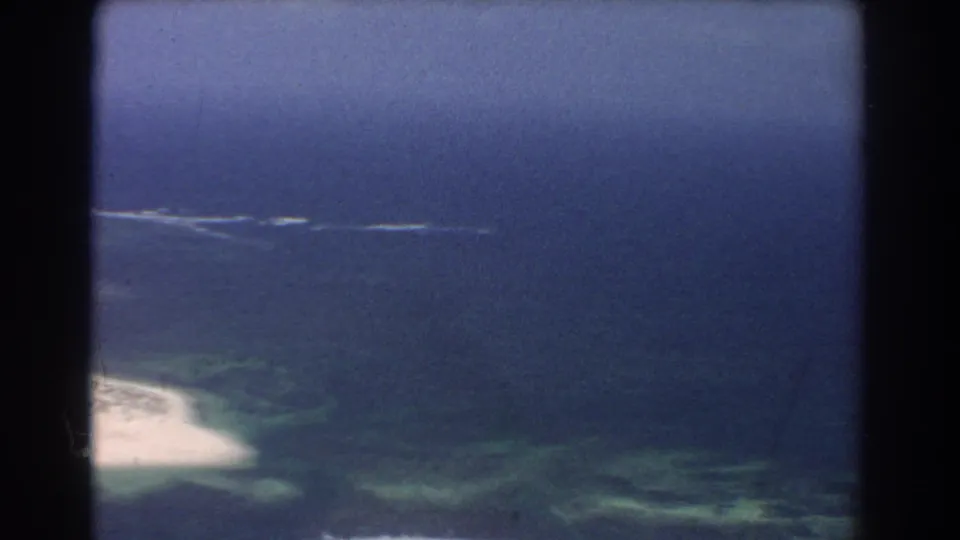 1967 Perth-area 8mm aerial frame showing blue ocean, a sandy edge, green-turquoise shallows, and dark film borders.