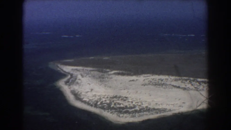 1967 Perth-area aerial 8mm frame showing a sandy coastal island edge, low vegetation, surf lines, and deep blue water.