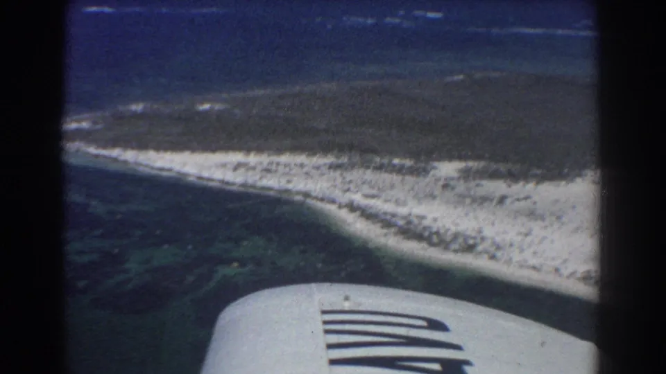 1967 aerial home-movie frame showing a curved coral coast, shallow turquoise water, and the edge of a plane wing.