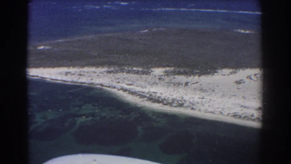 1967 8mm aerial view near Perth, Australia, showing a narrow sandy island edge, lagoon shallows, darker sea, and part of the aircraft frame.
