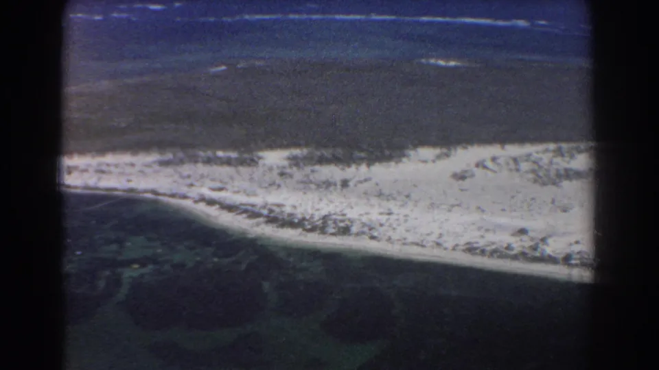 1967 8mm airplane-view footage near Perth, Australia, with a pale sandy shoreline dividing lagoon shallows from deeper blue water.