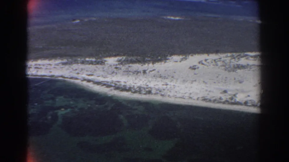 1967 8mm aerial view near Perth, Australia, looking down on a long pale beach, lagoon water, reef formations, and the lower edge of the aircraft.