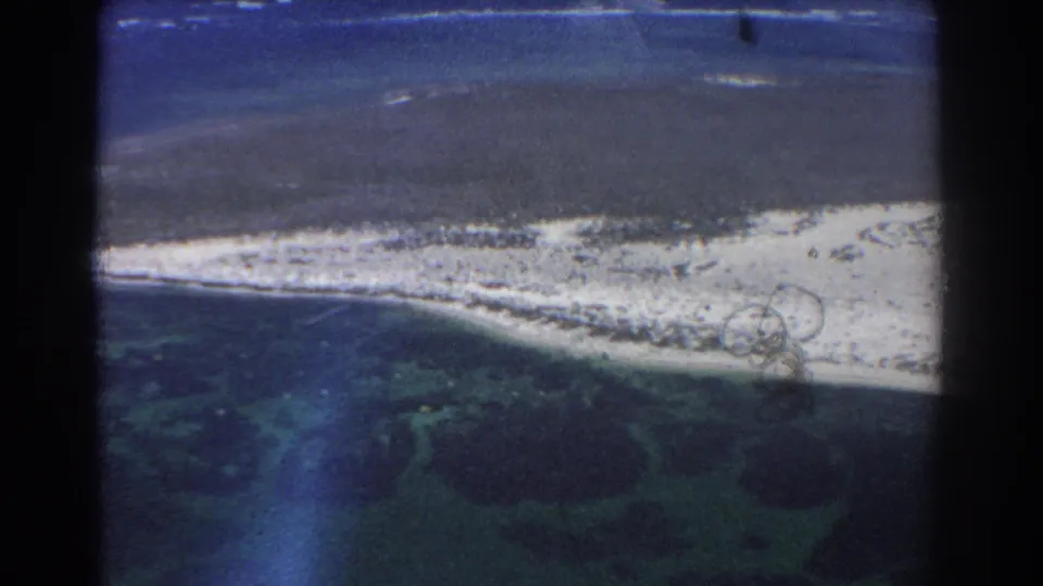 1967 8mm aerial coastal view near Perth, Australia, with turquoise lagoon patches, a pale beach line, darker ocean, and visible frame-edge artifacts.