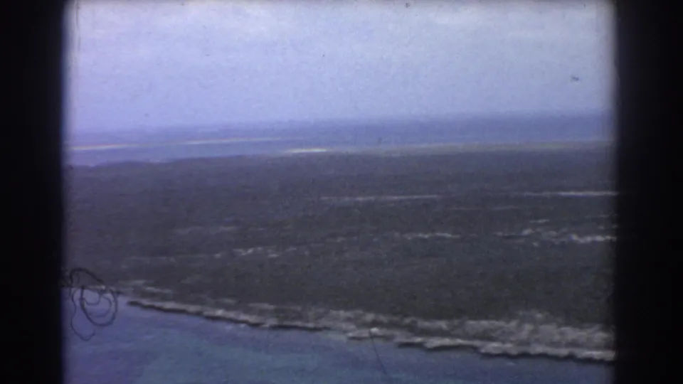 1967 home-movie aerial frame near Perth showing marsh, surf-marked shoreline, shallow water, and visible film wear.