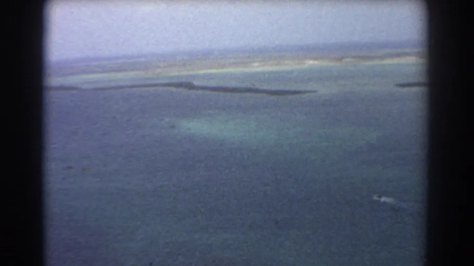 Aerial 8mm view near Perth showing blue ocean, pale shallow-water bands, a narrow shoreline, and aged film texture.