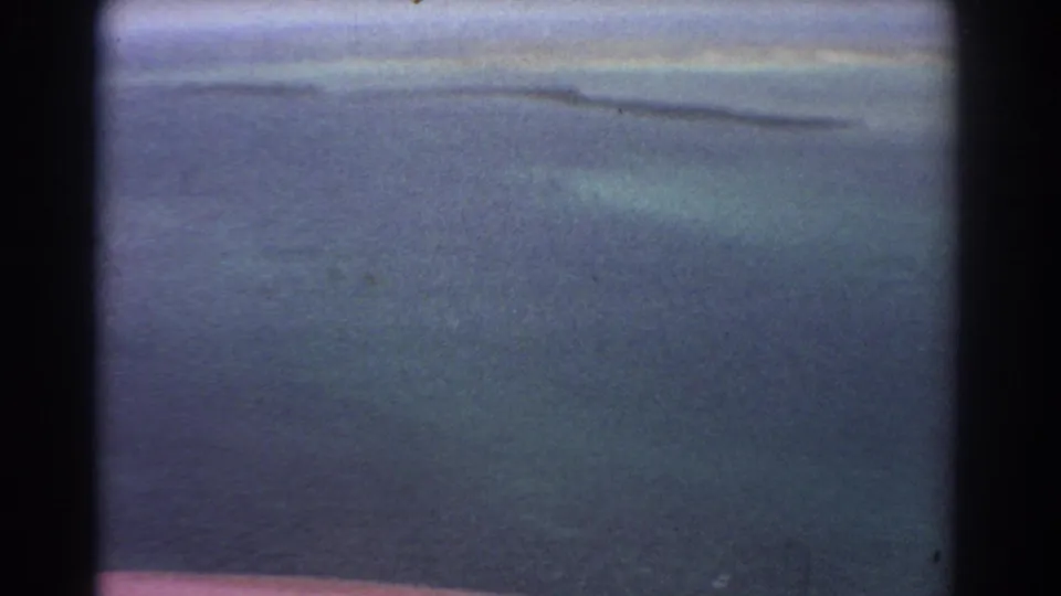 1967 8mm aerial view near Perth showing blue-green coastal water, distant sandbars, and a hazy horizon before the island enters frame.