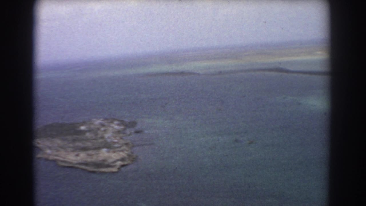 1967 8mm aerial frame near Perth with a small rugged island below, surrounded by blue and turquoise coastal water.