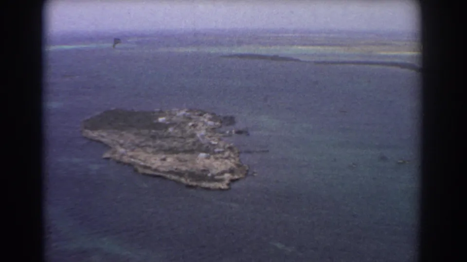 1967 8mm aerial flyby near Perth showing a small rugged island, dark wooded areas, pale shoreline, and surrounding blue-green water.