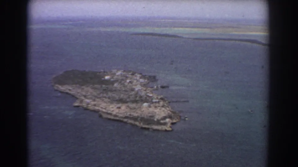 1967 8mm aerial view near Perth with the small island centered against turquoise shallows, deeper water, and a hazy distant coast.