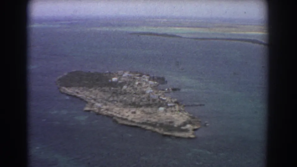 1967 8mm aerial home-movie frame showing a small island off Perth, Australia, with reef-toned shallows and scattered pale structures.