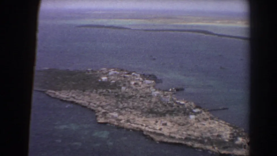 1967 8mm frame from a plane showing a closer view of the island, with rocky ground, sparse vegetation, and turquoise water nearby.